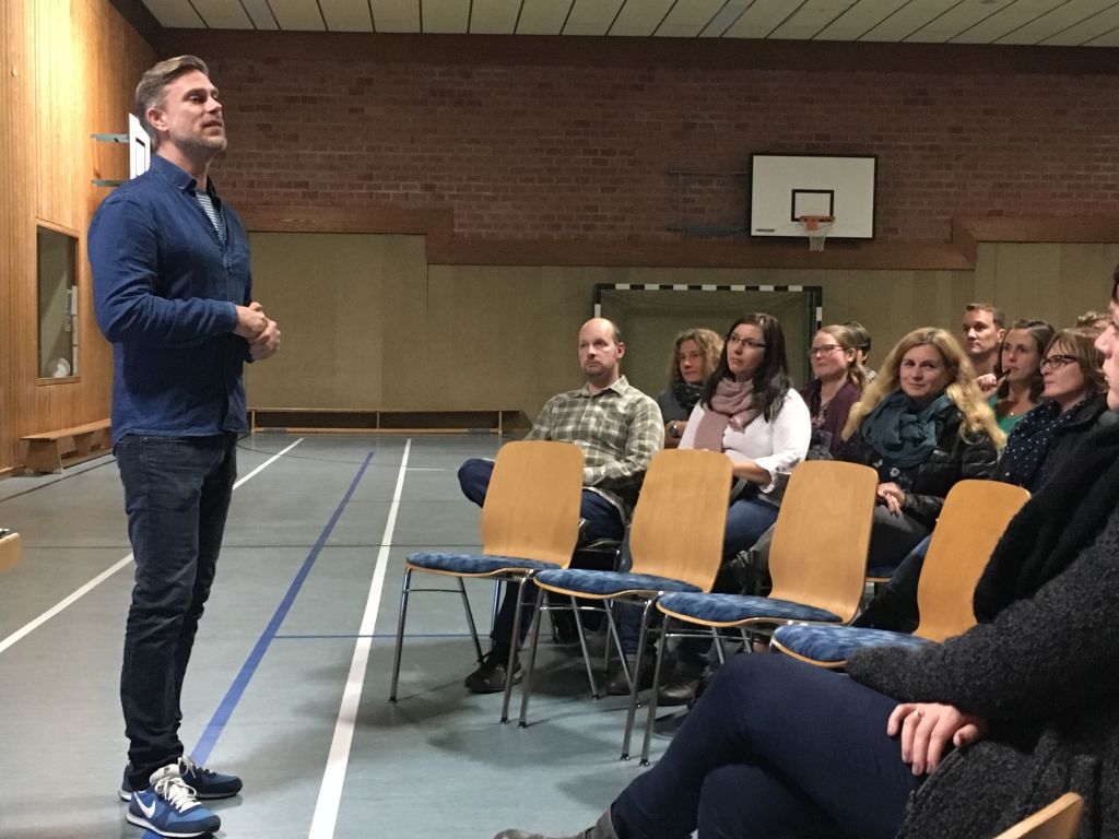 Graphik: Dozent hÃ¤lt Vortrag vor Lehrern und SchÃ¼lern in der Turnhalle